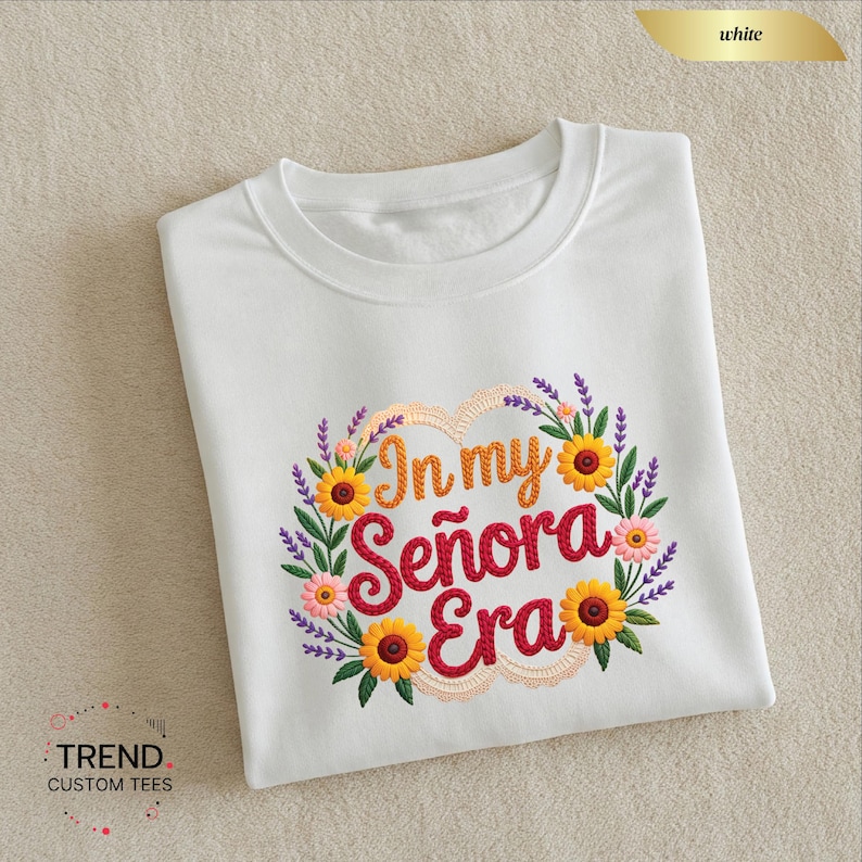 Op de afbeelding: Wit T-shirt met een bloemenontwerp en de tekst "In my Se&ntilde;ora Era" in een hartvormige krans. De krans bevat zonnebloemen, roze bloemen en paarse lavendel. Het shirt is op een neutrale ondergrond gevouwen.