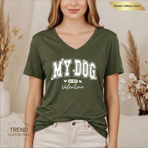 May include: Heather military green V-neck t-shirt with the text "MY DOG IS MY Valentine" in white. The text is in a collegiate style font with paw prints. The shirt is a solid color and has a relaxed fit.