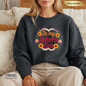 Op de afbeelding: Donkergrijze heather sweatshirt met de tekst "In my Se&ntilde;ora Era" in een bloemenkrans ontwerp. De krans bevat zonnebloemen, roze bloemen en paarse lavendel. De sweatshirt heeft een ronde hals.