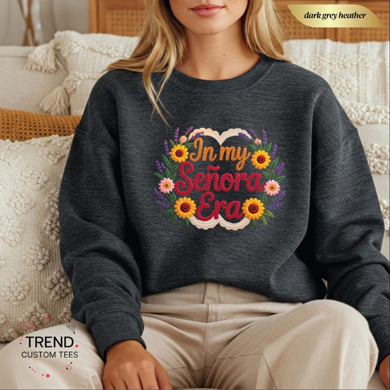 Op de afbeelding: Donkergrijze heather sweatshirt met de tekst "In my Se&ntilde;ora Era" in een bloemenkrans ontwerp. De krans bevat zonnebloemen, roze bloemen en paarse lavendel. De sweatshirt heeft een ronde hals.