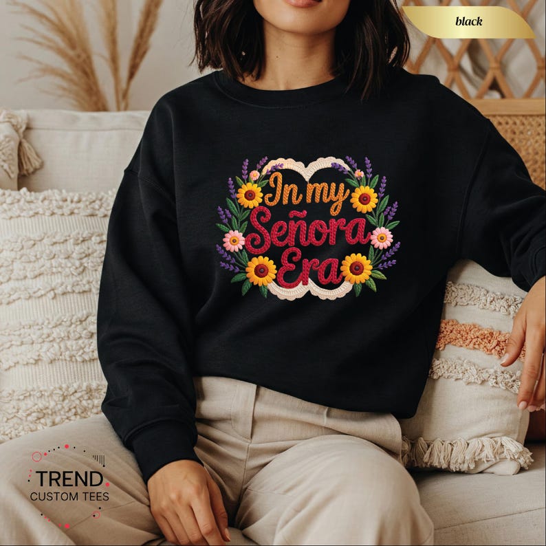Op de afbeelding: Zwarte sweatshirt met de tekst "In my Se&ntilde;ora Era" in een bloemenontwerp. Het ontwerp bevat zonnebloemen, lavendel en roze bloemen. De sweatshirt heeft een ronde hals.