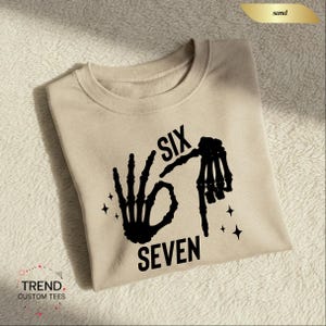 K&ouml;nnte beinhalten: Ein sandfarbenes Sweatshirt mit einer schwarzen Skeletthand-Grafik, die die Zahl sechs bildet, und dem Wort "SEVEN" darunter. Das Wort "SIX" steht &uuml;ber der Hand. Das Sweatshirt hat einen Rundhalsausschnitt.