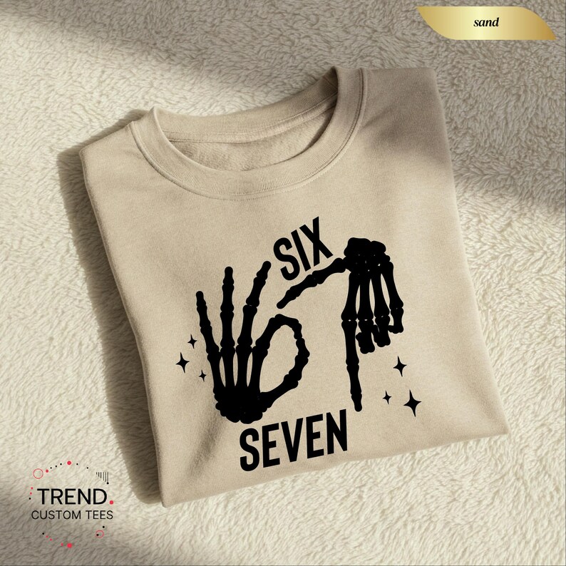 K&ouml;nnte beinhalten: Ein sandfarbenes Sweatshirt mit einer schwarzen Skeletthand-Grafik, die die Zahl sechs bildet, und dem Wort "SEVEN" darunter. Das Wort "SIX" steht &uuml;ber der Hand. Das Sweatshirt hat einen Rundhalsausschnitt.