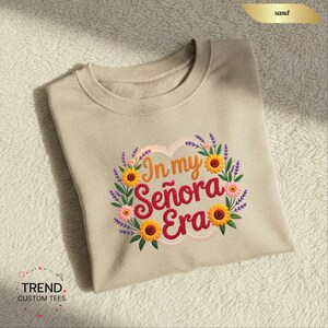 Op de afbeelding: Zandkleurige sweatshirt met geborduurd bloemmotief. Het ontwerp toont zonnebloemen, lavendel en de tekst "In my Se&ntilde;ora Era" in een hartvormige krans. De sweatshirt heeft een ronde hals en lange mouwen.