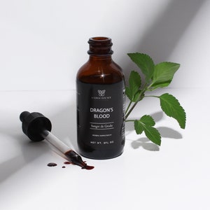 May include: A brown glass bottle with a dropper, labeled "Dragon's Blood Sangre de Grado Herbal Supplement, Net Wt. 2 Fl Oz." The bottle is sitting on a white surface with a green leaf.
