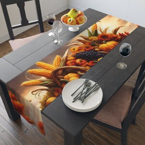 May include: A fall-themed table runner with a cornucopia overflowing with autumn harvest vegetables, fruits, and corn. The runner is set on a dark wood table with a white plate and silverware.