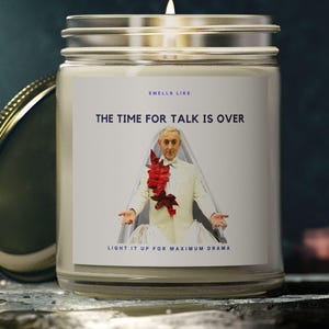 Puede incluir: Una vela blanca con una tapa dorada. La vela tiene una etiqueta que dice "Smells Like The Time For Talk Is Over" y presenta a un hombre con un traje blanco y una flor roja en el pecho. La etiqueta también dice "Light it up for maximum drama."