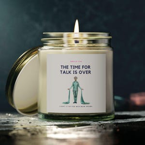 Puede incluir: Un tarro de vidrio para velas con tapa dorada. La vela está encendida y el tarro tiene una etiqueta blanca con el texto "THE TIME FOR TALK IS OVER" y una ilustración de dibujos animados de una persona con una túnica verde.