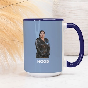 Puede incluir: Una taza de cerámica blanca con un borde y un asa azules. La taza presenta una foto de una mujer con una chaqueta de cuero negra y la palabra "MOOD" impresa debajo de la imagen.