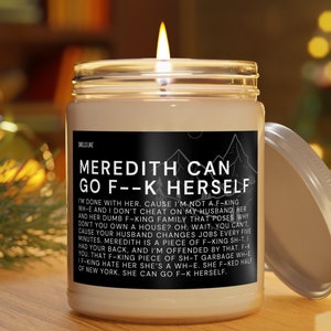 May include: A white candle jar with a black label that reads "Meredith Can Go F--K Herself". The label has a mountain range graphic and the text is in all caps. The candle is lit and the jar is sitting on a wooden surface.