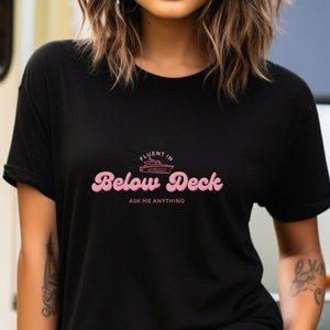 Op de afbeelding: Zwart t-shirt met de roze tekst "Fluent in Below Deck, Ask Me Anything". Het ontwerp bevat een kleine bootafbeelding boven de woorden "Below Deck".