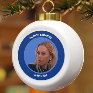 Puede incluir: Adorno navideño blanco con un círculo azul que presenta el rostro de una mujer y el texto "Sutton Stracke Name 'Em".