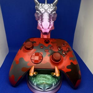 May include: A red and black camouflage Xbox controller is displayed on a 3D printed dragon-themed stand. The stand is silver and pink with a green and blue base.