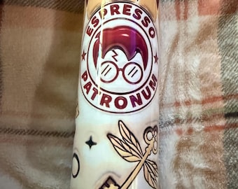 May include: A cylindrical tumbler with a maroon and gold geometric pattern at the top and bottom. The tumbler features the text "Espresso Patronum" around a graphic of a wizard's silhouette with glasses. Additional graphics include a key and a dragonfly.
