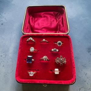 May include: A square, hinged jewelry box with a red velvet interior containing nine rings. The collection includes various metal bands featuring gemstones such as a large rectangular blue stone, a cluster of red stones, and several clear diamonds.