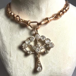 Op de afbeelding: Een vintage goudkleurige ketting met een decoratieve hanger. De hanger heeft een waaiervormig ontwerp versierd met talrijke heldere, sprankelende edelstenen. De ketting heeft een dikke ketting en een sluiting.