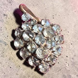 Op de afbeelding: Een clusterhanger met talloze ronde, gefacetteerde heldere stenen in een metalen zetting. De stenen zijn gerangschikt in een dichte, ronde formatie met een enkele lus aan de bovenkant voor bevestiging, wat een reflecterend oppervlak toont.