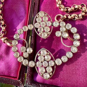 Può includere: Un set antico di collana e spilla color oro. La spilla presenta due sezioni ovali con pietre preziose rotonde e trasparenti. La collana ha un design a maglie. Il set è esposto in una scatola foderata di velluto viola.