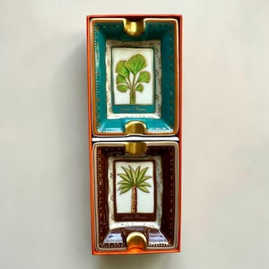 Op de afbeelding: Twee rechthoekige keramische asbakken in een oranje doos. De bovenste asbak is turquoise met een gouden rand en een groene plantenillustratie. De onderste asbak is bruin met een gouden rand en een palmboomillustratie. De tekst "palmier d'Hawaii" en "palmier Phoenix" is zichtbaar.
