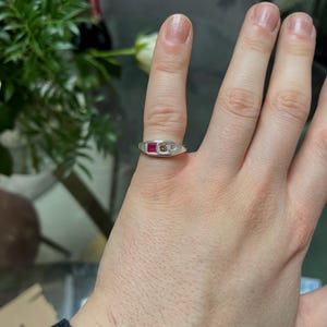 Pode incluir: Um anel de prata com uma pedra vermelha retangular, uma pedra castanha redonda e uma pequena pedra transparente. O anel est&aacute; num dedo, com um fundo desfocado de flores e uma mesa.