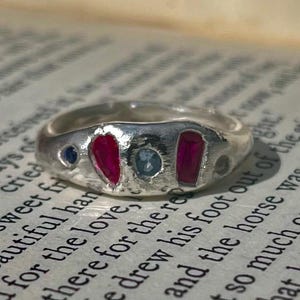 Könnte beinhalten: Ein silberner Ring mit unregelmäßigem Band, besetzt mit einem herzförmigen Rubin, einem rechteckigen Rubin und zwei kleineren blauen Edelsteinen. Der Ring wird auf einer Vintage-Buchseite gezeigt.