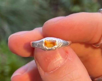 May include: A silver ring with a rectangular, orange gemstone. The ring has a textured, organic design, and the gemstone is set in the center. The ring is held between fingers, showcasing the jewelry's details and color.