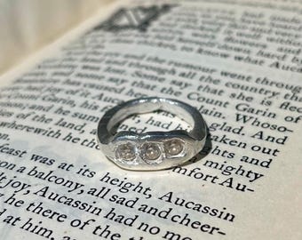 May include: A silver ring with three small, clear stones set in a row. The ring is on a page of an open book with text. The ring has a textured, slightly uneven surface.