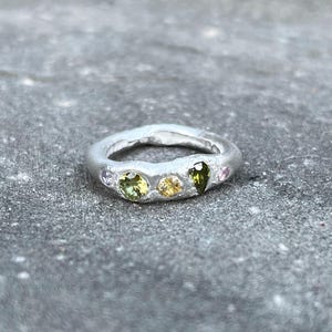 Op de afbeelding: Een zilveren ring met een getextureerde band en vijf kleine edelstenen. De edelstenen zijn groen, geel en roze.