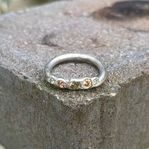 May include: A silver ring with five small, colorful gemstones set in a row. The ring is on a gray, textured surface.