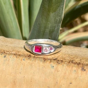 May include: A silver ring featuring a square-cut vibrant red gemstone set alongside two smaller round stones, one light pink and one deep pink. The band has a smooth, polished finish and a rounded profile, showcasing a cluster of three distinct gems.