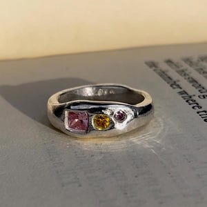 May include: A silver ring with three gemstones. The ring has a pink square-cut stone, a yellow round stone, and a small purple round stone. The ring is set against the backdrop of an open book.