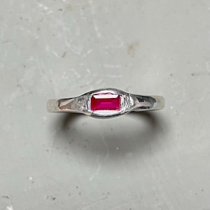 Op de afbeelding: Een zilveren ring met een rechthoekige, helderrode edelsteen. De band is glad en gepolijst, met een getextureerde zetting rond de edelsteen. De ring is een eenvoudig maar elegant sieraad.