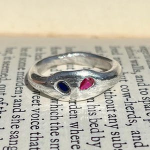Könnte beinhalten: Ein silberner Ring mit strukturiertem Band, mit einem tropfenförmigen, tiefrosa Edelstein und einem tropfenförmigen, dunkelblauen Edelstein. Der Ring ist vor einem Hintergrund aus gedrucktem Text platziert.