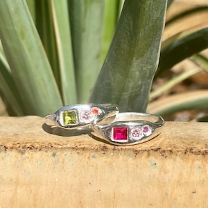 May include: Two silver rings featuring multi-coloured gemstones. The first ring has a square green stone and two small round pink stones. The second ring has a square red stone and two small round pink stones. Both rings have a smooth, polished band.