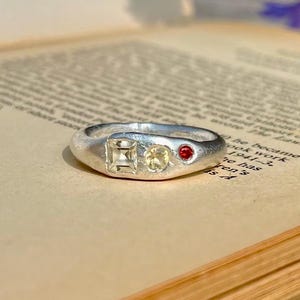 Puede incluir: Un anillo de plata con tres piedras preciosas: una cuadrada, una redonda y una pequeña piedra roja. El anillo está sobre una página de libro antiguo con texto visible. Las piedras son claras y el anillo tiene una banda lisa y redondeada.