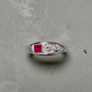 Può includere: Un anello d'argento con una pietra quadrata color rubino, una pietra rotonda color champagne e una piccola pietra rosa. L'anello ha una finitura liscia e lucida. L'anello è un gioiello.