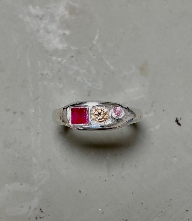Pode incluir: Um anel de prata com uma pedra quadrada de cor rubi, uma pedra redonda cor champanhe e uma pequena pedra rosa. O anel tem um acabamento liso e polido. O anel &eacute; uma joia.