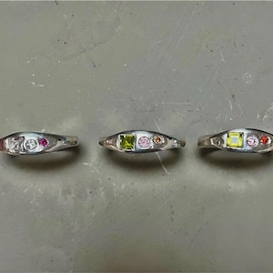 以下が含まれることがあります： それぞれがユニークな配置のカラフルな宝石がセットされた3つのシルバーリング。1つのリングは、正方形、丸、ピンクの宝石が特徴です。別のリングは、緑色の正方形、ピンクの丸、オレンジ色の丸い宝石が付いています。3番目のリングは、白い正方形、丸、ピンクの宝石が付いています。