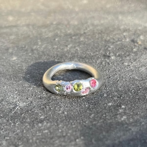 Pode incluir: Um anel de prata com uma banda texturizada e um grupo de pedras preciosas rosa e verdes.