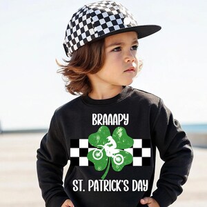 May include: A black sweatshirt with a green shamrock graphic and the text "BRAAAPY ST. PATRICK'S DAY". The shamrock features a checkered flag and a person riding a dirt bike. The model is wearing a black and white checkered cap.