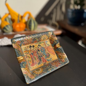 May include: Square decorative ashtray with an Asian-inspired design. The ashtray features a central panel depicting figures in traditional attire, surrounded by floral and geometric patterns in gold, blue, and red. The edges are trimmed with gold.