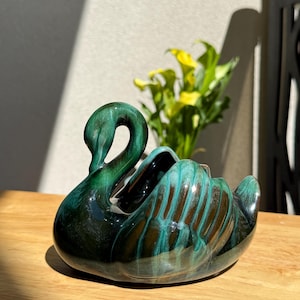 May include: A ceramic swan planter with a glossy, mottled green and teal glaze. The swan has a curved neck and open space for plants or decor. The planter sits on a wooden surface, with yellow flowers in the background.