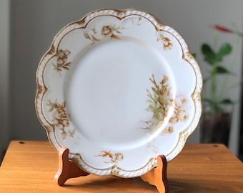 Peut inclure: Une assiette en porcelaine blanche avec un bord festonné et une bordure dorée. L'assiette présente un motif floral avec des roses brunes et vertes.