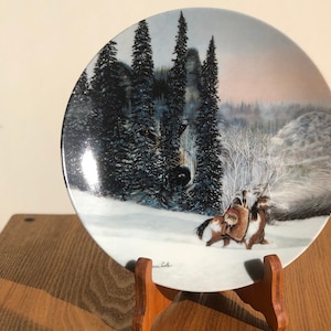 Puede incluir: Un plato de porcelana blanca con una escena de invierno que presenta un lobo asomándose entre los árboles, un paisaje nevado y dos figuras a caballo. El plato está sobre un soporte de madera.