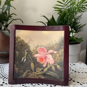 May include: A framed painting featuring a pink orchid and a hummingbird. The artwork is set against a backdrop of lush greenery and a soft, golden sky. The frame is a dark, rich brown, adding a classic touch to the floral scene.