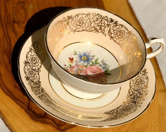 Puede incluir: Un delicado juego de taza y platillo de té con base rosa y blanca y ribete dorado. La taza presenta un diseño floral con flores rosas, azules y amarillas.