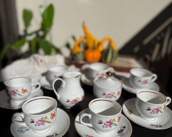 Könnte beinhalten: Ein weißes Porzellan-Teeservice mit floralen Mustern. Das Set umfasst Tassen, Untertassen, eine Milchkanne und eine Zuckerdose. Jedes Stück ist mit bunten Blumen und Goldrändern verziert, was eine klassische und elegante Ästhetik schafft.