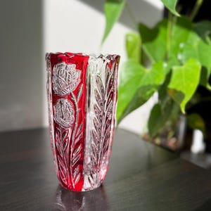 May include: A decorative glass vase with a red interior and detailed floral and leaf patterns. The vase is a clear, cylindrical shape with a flared rim. The carvings create a contrast between the red and clear glass.