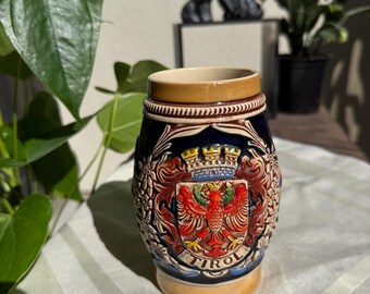 May include: A ceramic beer stein with a blue body, tan rim, and base. The stein features a detailed crest with a red eagle, a crown, and the word "TIROL". Floral and decorative elements surround the crest.
