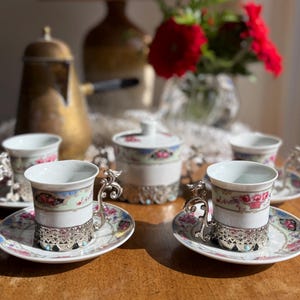 May include: Set of four porcelain teacups and saucers with floral designs and silver accents. The cups and saucers feature a delicate floral pattern in shades of pink, blue, and green. Each cup has a silver handle and a silver base. The saucers have a matching floral design and a silver rim.